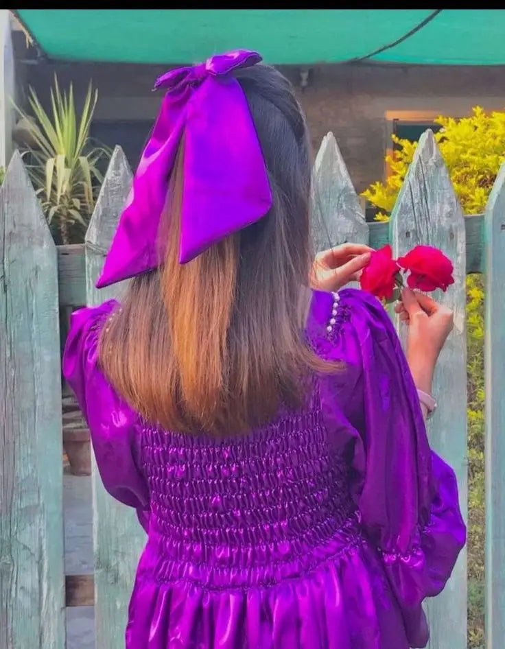 Person wearing a purple dress with a large bow in their hair, standing in front of a wooden fence.